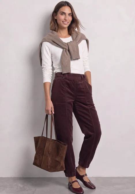 High Waist Barrel Leg Hose mit Elastikbund vintage ruby Frau in weißem Pullover, brauner Cordhose und passenden Accessoires mit großer Wildledertasche.