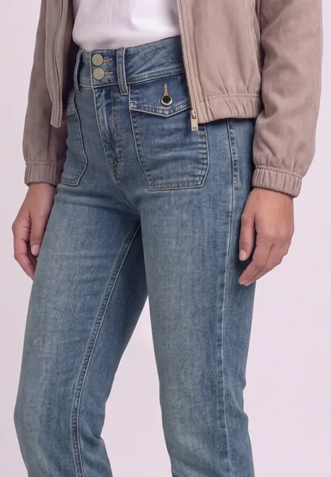 Person in heller Jacke und hoch taillierter blauer Jeans mit Zierknöpfen, sichtbar bis zu den Oberschenkeln.