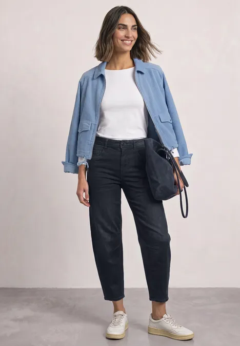 High Waist Barrel Leg Jeans im Loose Fit blue black soft wash Frau in hellblauer Jacke, weißem T-Shirt, dunklen Jeans und Sneakern mit navy Tasche vor neutralem Hintergrund.