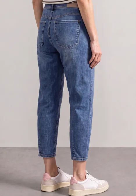 Rückenansicht einer Person in hochtaillierten blauen Jeans und weißen Sneakern vor neutralem Hintergrund.