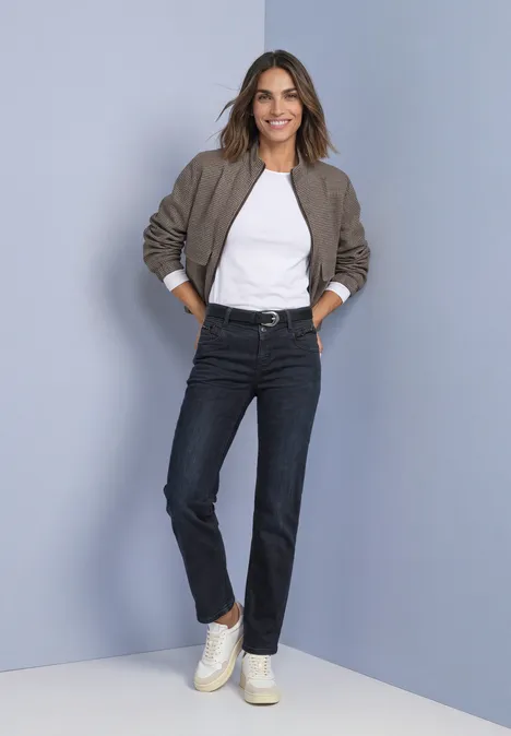 Mid Waist Straight leg Jeans im Casual Fit blue black soft wash Frau in weißem T-Shirt, Bomberjacke, dunklen Jeans und Sneakern vor grauem Hintergrund.