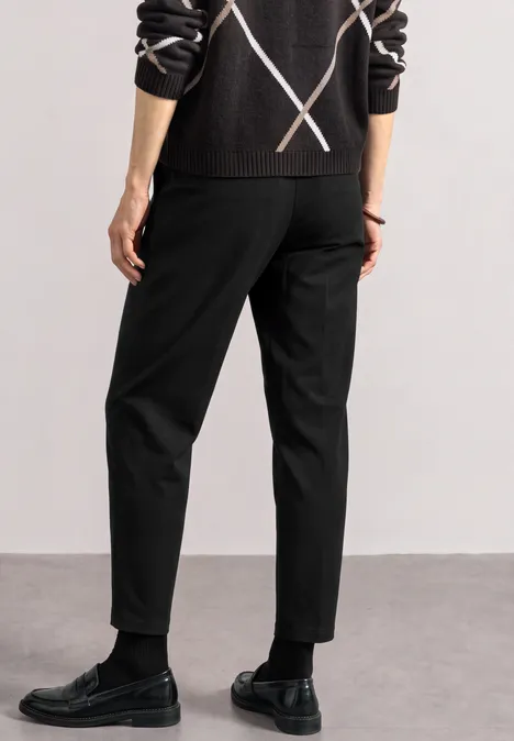 Person von hinten in schwarzen Hosen, schwarzen Lederschuhen und dunklem Pullover mit X-Muster.