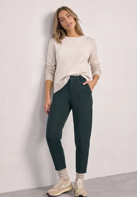 Eine Frau steht vor einer hellen Wand, trägt beigen Pullover, grüne Hose und helle Schuhe, lächelnd.