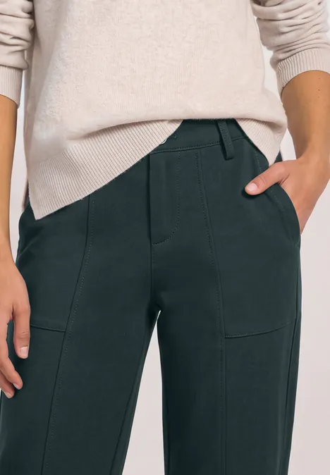 Weibliche Person in hellem Pullover und dunkler Hose, Hände in den Taschen, Close-up auf Oberkörper und Beine.