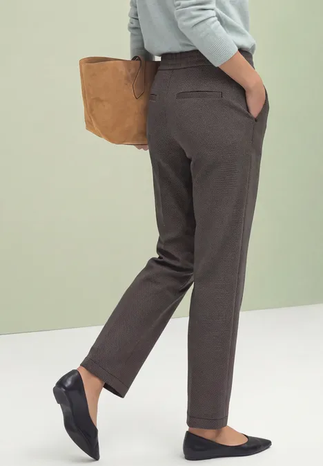 Femme en pantalon gris-brun droit, ballerines noires, tenant un sac marron.