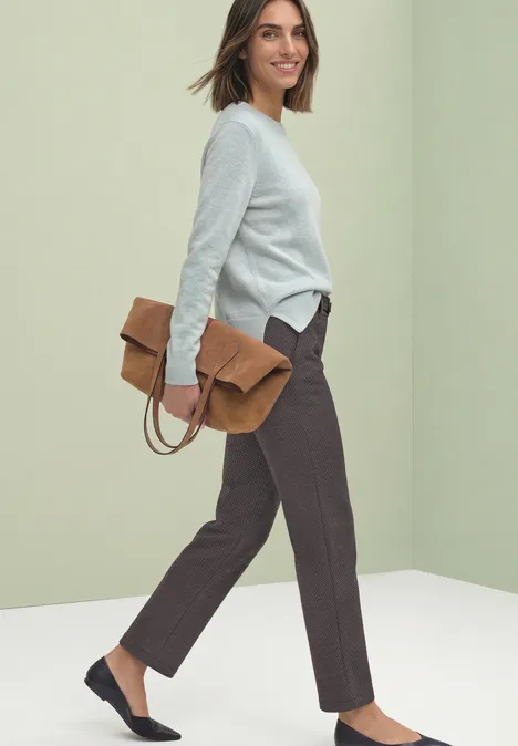 Une femme porte un pull gris clair-bleu, un pantalon marron et des ballerines bleu nuit, tenant une pochette marron.