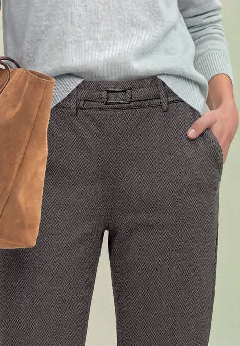 Gros plan sur pantalon gris texturé avec ceinture, main dans la poche, pull bleu clair, sac en daim brun.