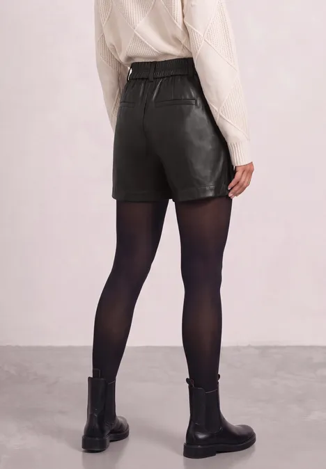 Rückansicht einer Person mit beigem Strickpullover, schwarzen Leder-Shorts, schwarzen Strumpfhosen und schwarzen Boots.