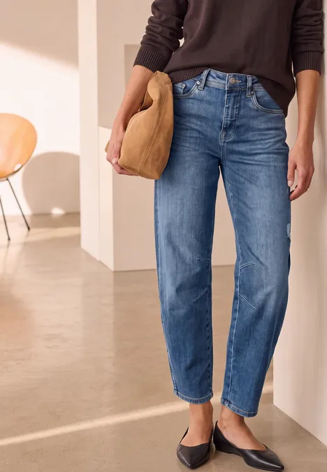 Frau trägt gerade geschnittene blaue Jeans, schwarze Ballerinas und beige Clutch vor minimalistischem Interieur.