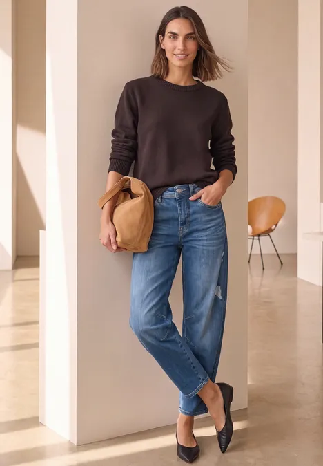 Frau in braunem Pullover und Jeans mit Strohhut in heller, minimalistischer Umgebung.