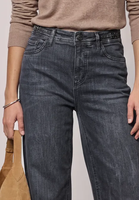 Jean à jambes larges avec des détails au niveau de la ceinture Steel Grey Washed Femme portant un jean cargo noir taille haute, un top beige à manches longues et tenant un sac marron.