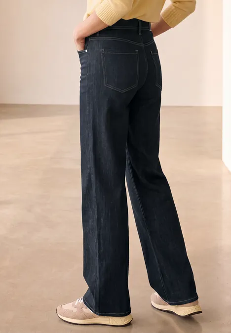 Wide Leg Jeans mit details am Bund Rinse Washed Rückansicht einer Person in dunklen, weiten Jeans und hellen Sneakers vor neutralem Hintergrund.