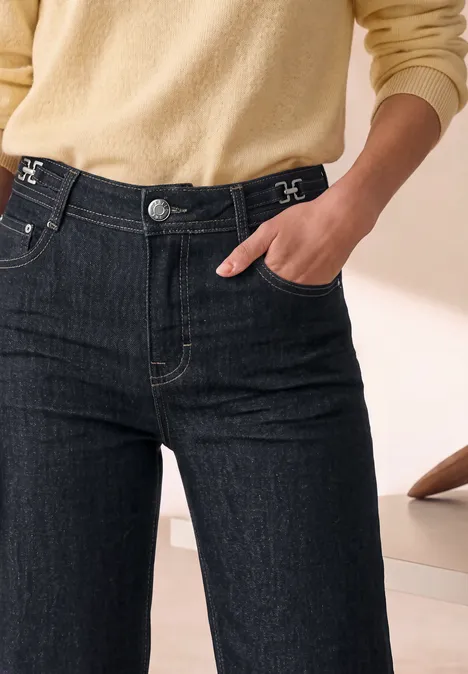 Wide Leg Jeans mit details am Bund Rinse Washed Person trägt dunkle High-Waist-Jeans und hellbeigen Pullover, Hand in Tasche, minimalistische Wohnraum-Kulisse.