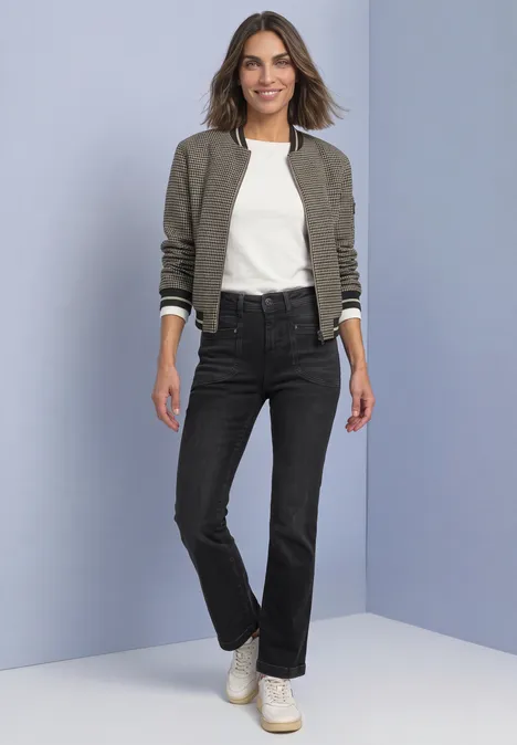 Jean bootcut taille haute, coupe slim Black Washed Frau trägt olivgrüne Bomberjacke, weißes Shirt, schwarze Jeans und Sneakers, steht vor blauem Hintergrund.