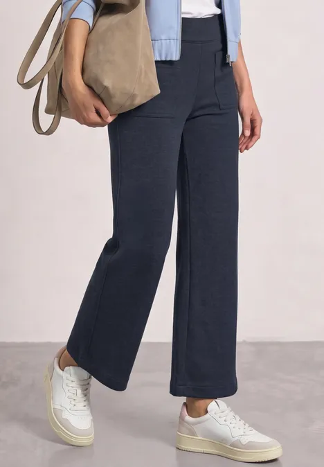 Wide Leg Hose mit aufgesetzten Taschen darkest blue mel. Untersicht einer Person in dunkelblauen, weiten Hosen und weißen Sneakers, mit beiger Tasche.