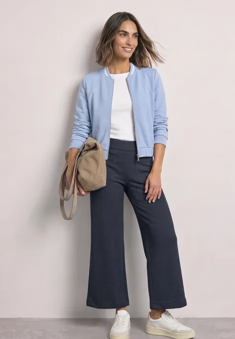 Wide Leg Hose mit aufgesetzten Taschen darkest blue mel. Eine lächelnde Frau trägt eine hellblaue Jacke, weißes T-Shirt, dunkle Schlaghose, weiße Sneakers und eine beige Tasche.