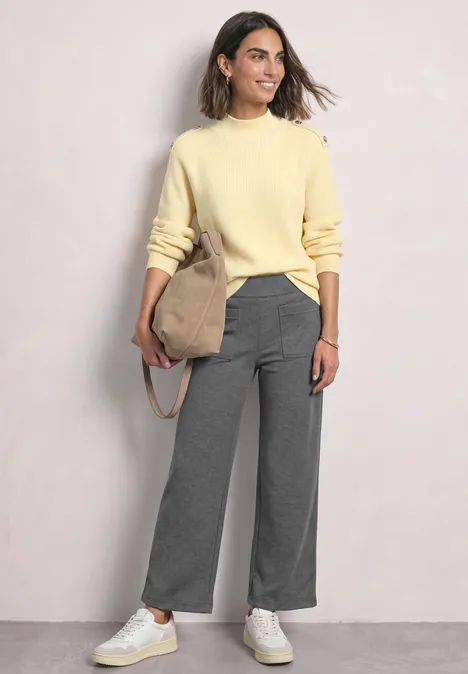 Eine Frau trägt hellgelben Pullover, graue weite Hose, weiße Sneaker und eine Beuteltasche.