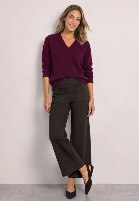 Een vrouw in een bordeauxrode V-hals trui, zwarte wijde 7/8 broek en zwarte ballerina's voor een lichte achtergrond