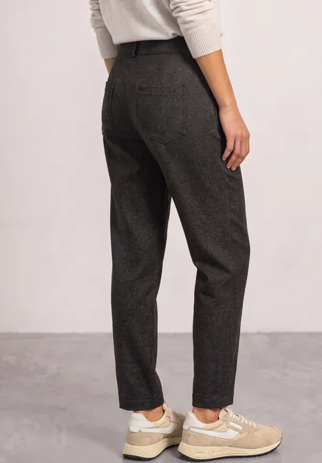 Vrouw van achteren in donkergrijze stoffen broek en beige sneakers tegen neutrale achtergrond.