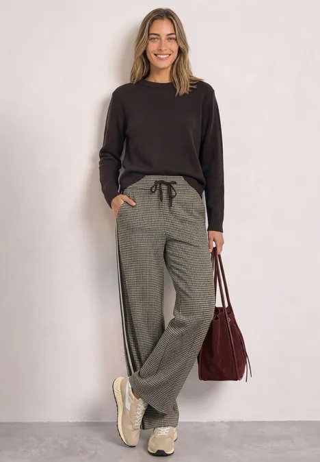Lächelnde Frau in schwarzem Pullover, grauen Jogginghosen und Sneakers hält rote Tasche in lockerer Pose.