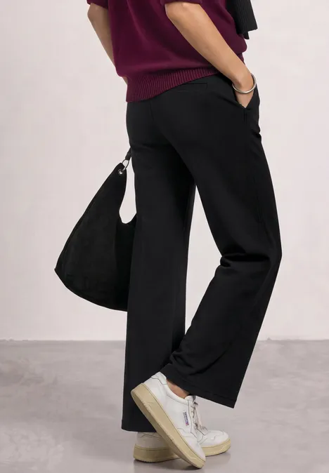 Zijaanzicht van een persoon in zwarte wijde broek, witte sneakers, bordeaux trui en schoudertas.