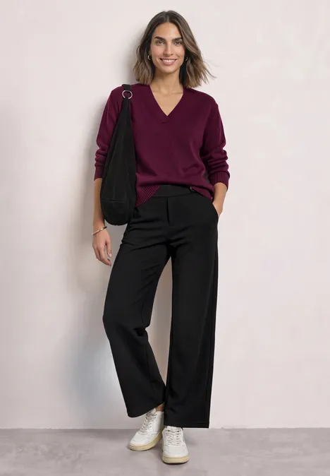 Vrouw leunt tegen muur, draagt bordeauxrode V-hals trui, zwarte wijdvallende broek, witte sneakers en zwarte tas.