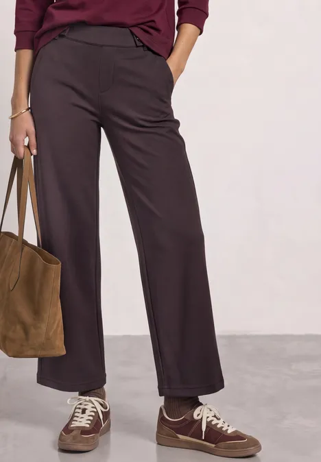Eine Person trägt weite braune Hosen, braune Sneaker und hält eine braune Veloursledertasche.
