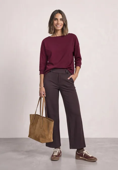 Frau in burgunderfarbenem Pullover, weitem Hosenrock und Ledertasche vor neutralem Hintergrund.