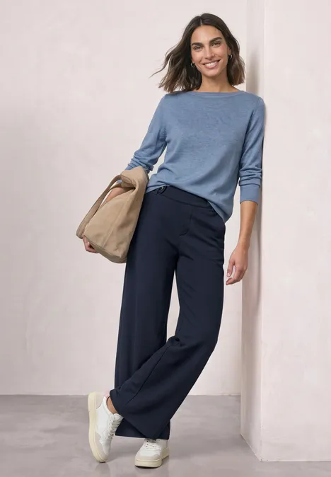 Vrouw in blauwe trui en losse marineblauwe broek met witte sneakers en beige tas, nonchalant tegen muur geleund.