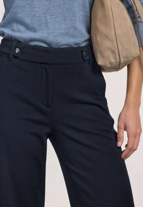 Close-up van een taille met donkerblauwe broek, lichtblauw shirt en beige handtas.