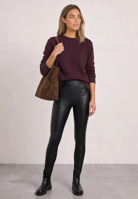 Eine Frau steht in schwarzer Lederhose, schwarzen Stiefeln, bordeauxrotem Pullover und brauner Tasche.