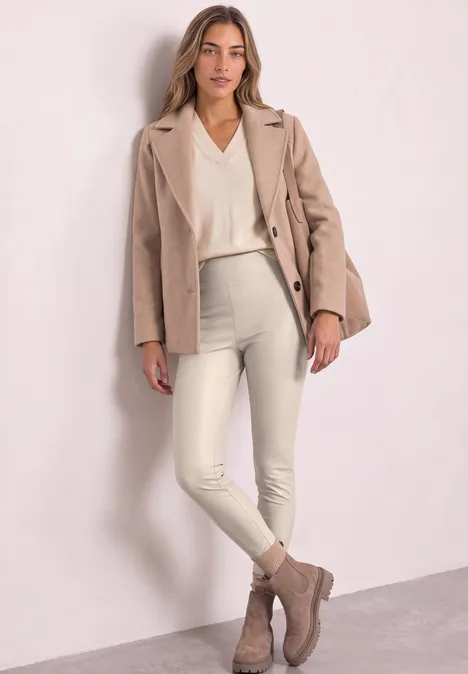 Frau in beigem Blazer, cremeweißem Pullover, weißen Hosen und taupefarbenen Stiefeletten vor heller Wand.