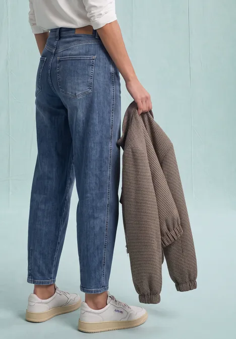 High Waist Barrel Leg Jeans im Loose Fit Medium Blue Destory Washed Person von hinten in lockeren Jeans und weißen Sneakern, hält eine graubraune Jacke in der Hand.
