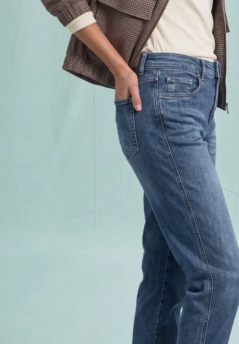 High Waist Barrel Leg Jeans im Loose Fit Medium Blue Destory Washed Eine Person trägt blaue Jeans und eine braune Jacke, steht seitlich mit Hand in Tasche.