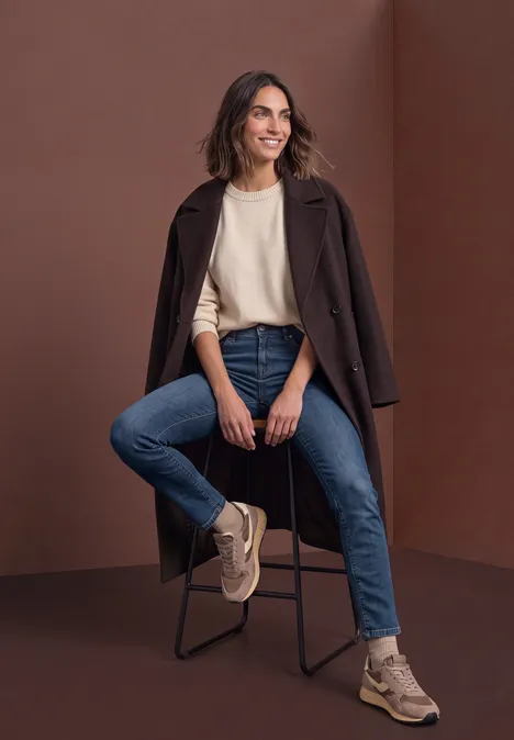 Frau sitzt lässig auf einem Hocker, trägt Jeans, hellen Pulli, beigen Mantel über den Schultern und Sneaker.