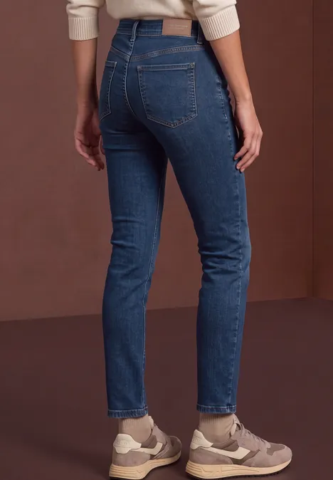 Rückansicht einer Person in blauen Jeans und Sneakern vor braunem Hintergrund.