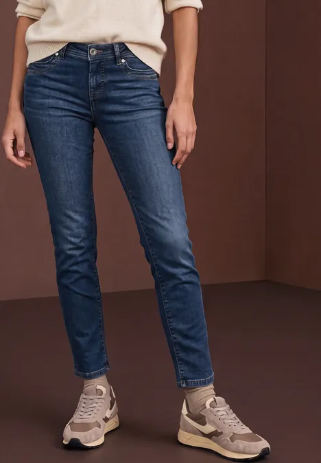 Frau trägt blaue Jeans und Sneaker vor braunem Hintergrund.