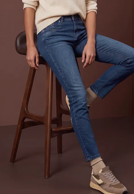 Person sitzt auf hohem Holzstuhl, trägt enge blaue Jeans und beige Sneaker.
