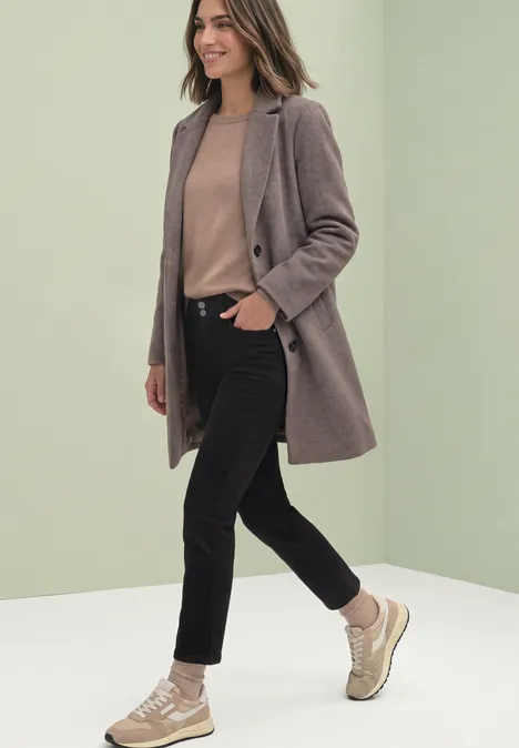 Frau in modernem Mantel, beigem Pullover, schwarzen Jeans und Sneakers vor hellgrünem Hintergrund.
