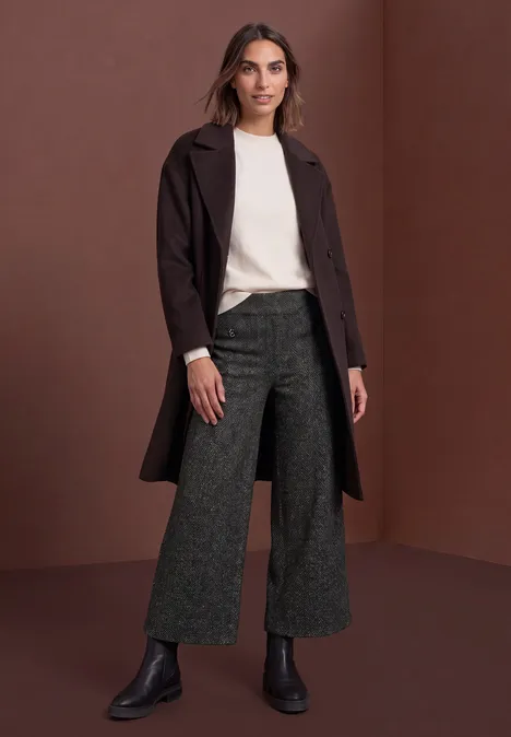 Vrouw met een crème trui, donkerbruine mantel, wijde grijze broek en zwarte boots voor een bruine achtergrond.
