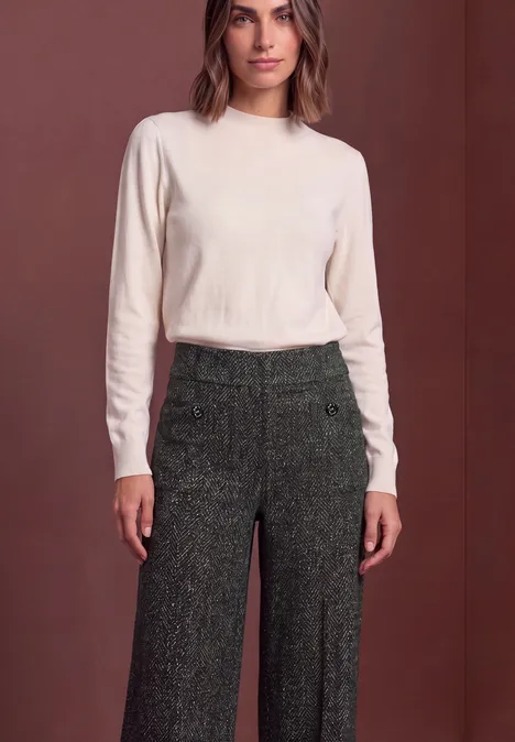Vrouw in een crèmekleurig langemouwen shirt en wijde, donkergroene tweedbroek voor een effen achtergrond.