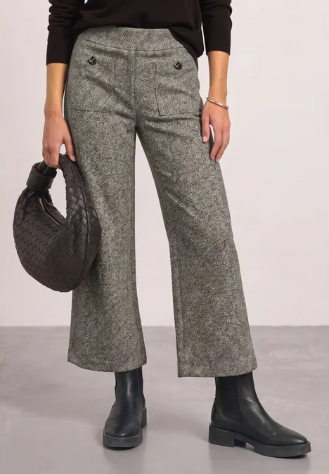 Grijze, wijd uitlopende broek met knoopdetail, zwarte gevlochten handtas en zwarte laarzen.