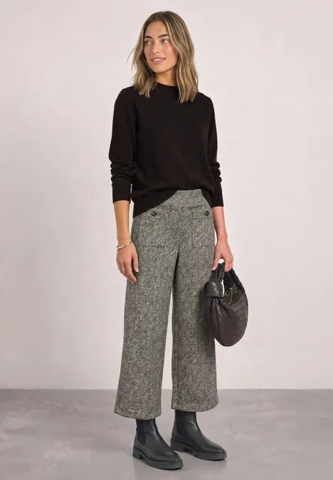 Vrouw in zwarte trui, grijze culotte, zwarte laarzen en met zwarte tas voor een neutrale achtergrond.