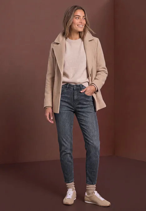 Femme en manteau beige, haut blanc, jean foncé et baskets devant un fond marron.