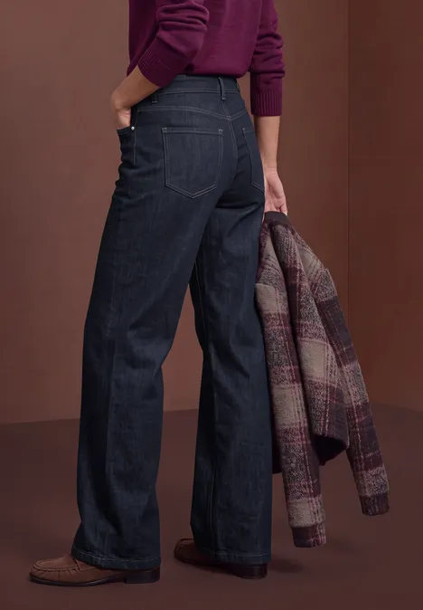 Jeans met High Waist en Wide Leg pijpen in een Loose Fit pasvorm Rinse Washed Eine von hinten fotografierte Frau trägt dunkle, weite Jeans, braune Schuhe und hält eine karierte Jacke.