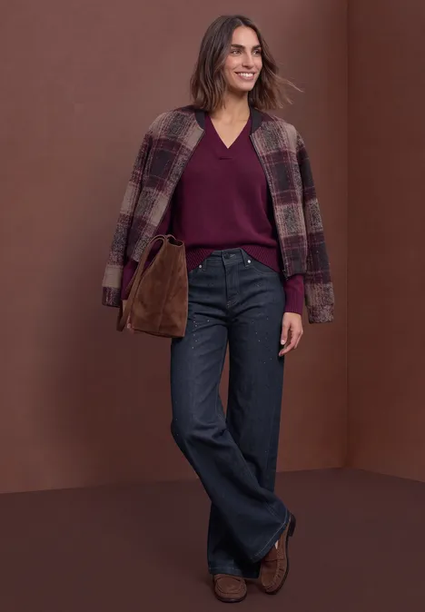 Jeans met High Waist en Wide Leg pijpen in een Loose Fit pasvorm Rinse Washed Frau trägt karierten Mantel über den Schultern, Burgunderpullover, weite Jeans und braune Schuhe vor braunem Hintergrund.