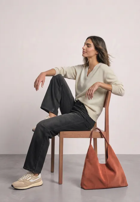 Mid Waist Wide Leg Jeans im Loose Fit Black Washed Eine Frau sitzt übereinandergeschlagen auf einem Holzstuhl, trägt beiges Oberteil, schwarze Hose, Sneaker und rote Tasche.