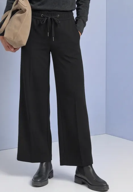 Mid Waist Straight Leg Hose im Loose Fit Black Schwarze, weit geschnittene Hose mit Tunnelzug, schwarze Stiefel und beige Tasche vor hellblauem Hintergrund.