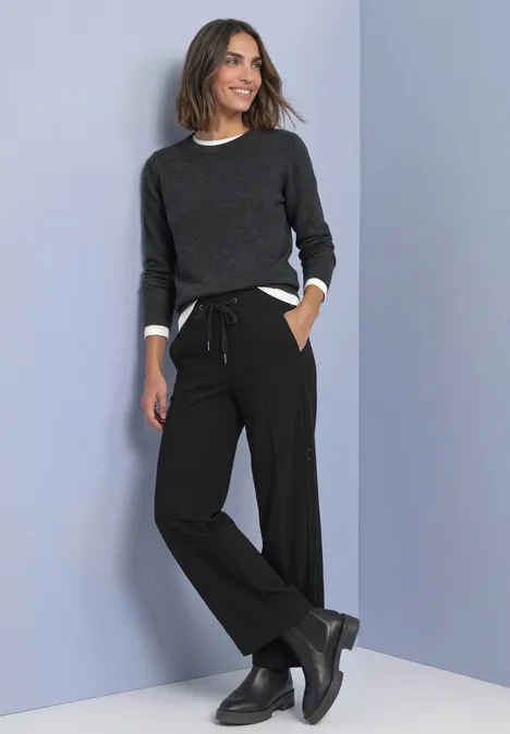 Mid Waist Straight Leg Hose im Loose Fit Black Frau in dunkelgrauem Pullover und weiten schwarzen Hosen mit Kordelzug und schwarzen Stiefeln vor hellem Hintergrund.
