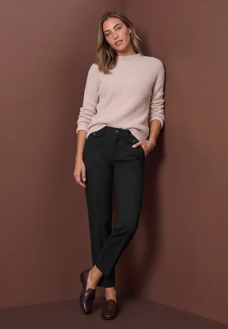 Une femme souriante en pull beige et pantalon noir pose devant un fond marron.